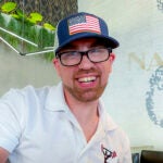 James Norris, a 37-year-old man, is pictured smiling. He has a short, sandy-colored beard, black-framed glasses, and a baseball cap with an American flag decal. He is wearing a white, collared short-sleeved shirt.