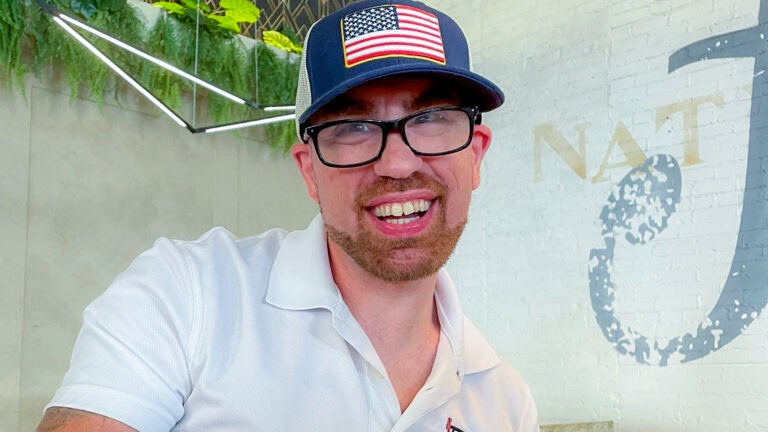 James Norris, a 37-year-old man, is pictured smiling. He has a short, sandy-colored beard, black-framed glasses, and a baseball cap with an American flag decal. He is wearing a white, collared short-sleeved shirt.