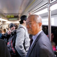 MBTA General Manager Phillip Eng is seen in the foreground on a crowded Green Line train. He is wearing a suit and smiling.