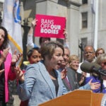 Gov. Maura Healey stands at a dais, wearing a light blue suit and standing in front of an excited crowd, where one person can be seen holding a sign that reads "bans off our bodies."