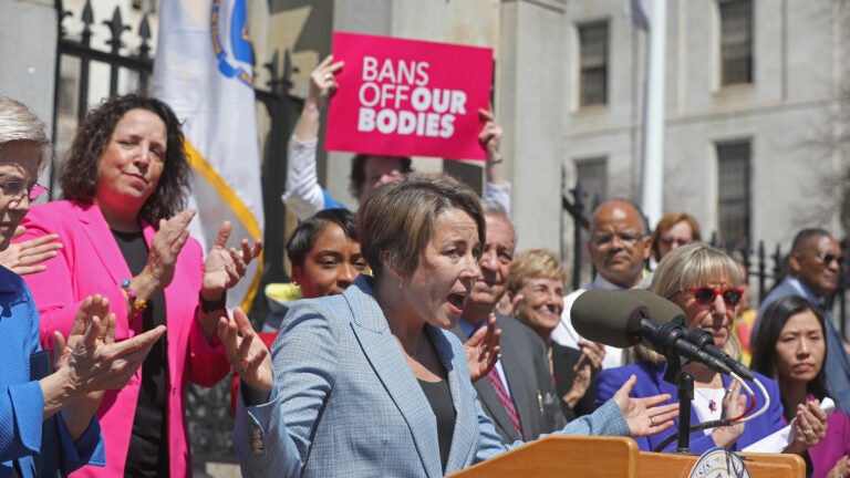 Gov. Maura Healey stands at a dais, wearing a light blue suit and standing in front of an excited crowd, where one person can be seen holding a sign that reads "bans off our bodies."