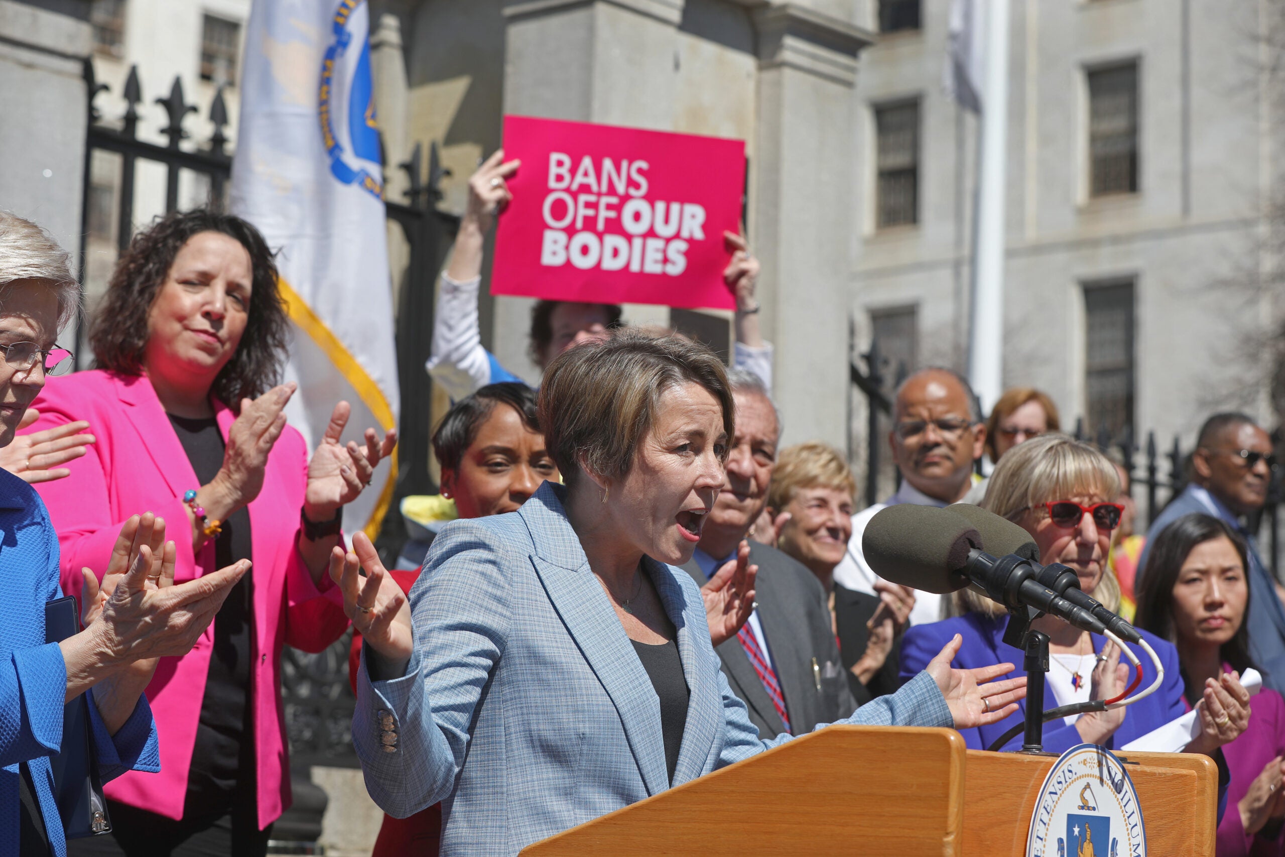 Gov. Maura Healey stands at a dais, wearing a light blue suit and standing in front of an excited crowd, where one person can be seen holding a sign that reads 