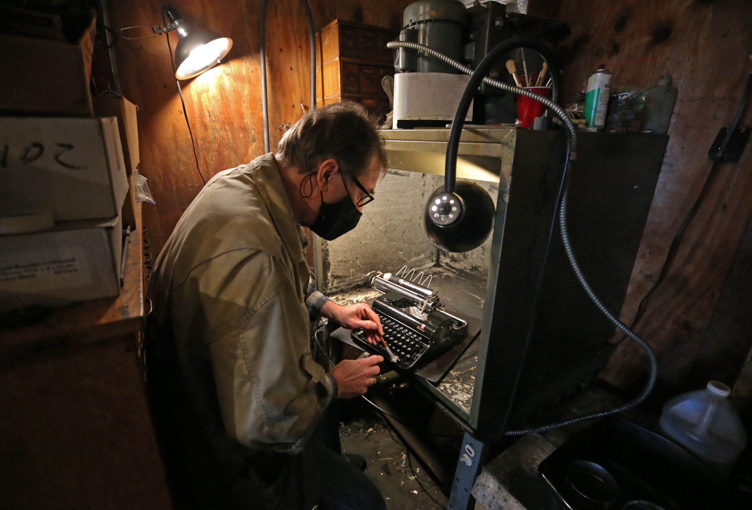 Cambridge Typewriter owner Tom Furrier hunches over a typewriter as he holds a tool to the machine. He is wearing a tan jacket, glasses, and a black face mask.