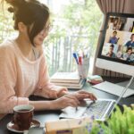 An Asian woman in a pink sweater works at a desk in a home office. There is a Zoom meeting on the screen, coffee in a brown mug on a saucer, a laptop, and a plant in the foreground.