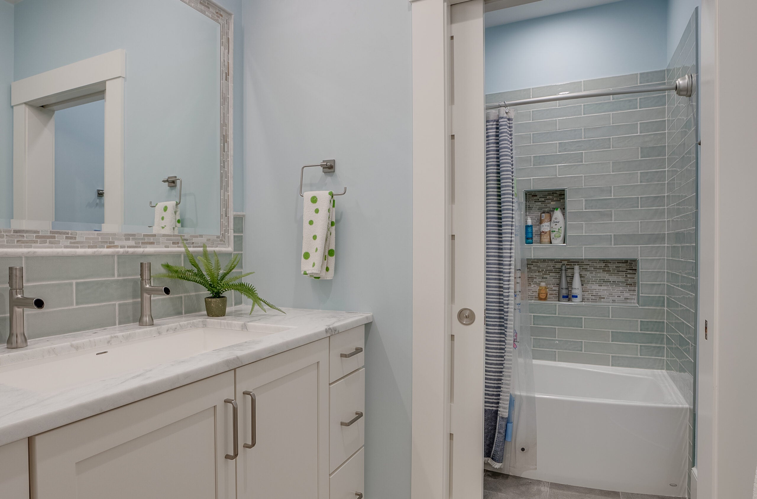 A bathroom with a trough sink, two faucets, and a pocket door that opens to reveal a tub/shower combination with a shower curtain and tile backsplash.
