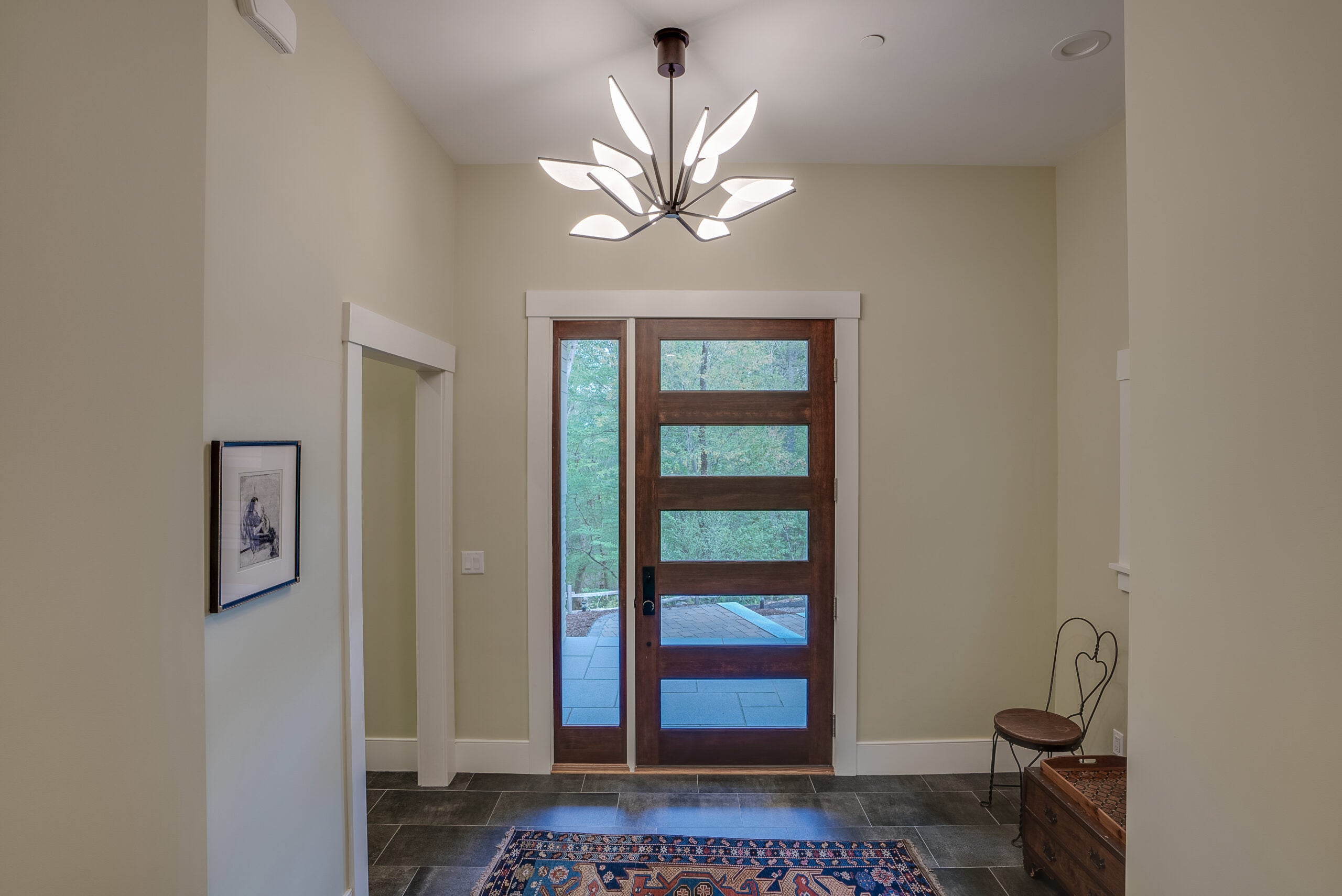 A six-panel wooden door with a sidelight in a foryer with a tile floor and a modern chandelier that looks like a starburst.