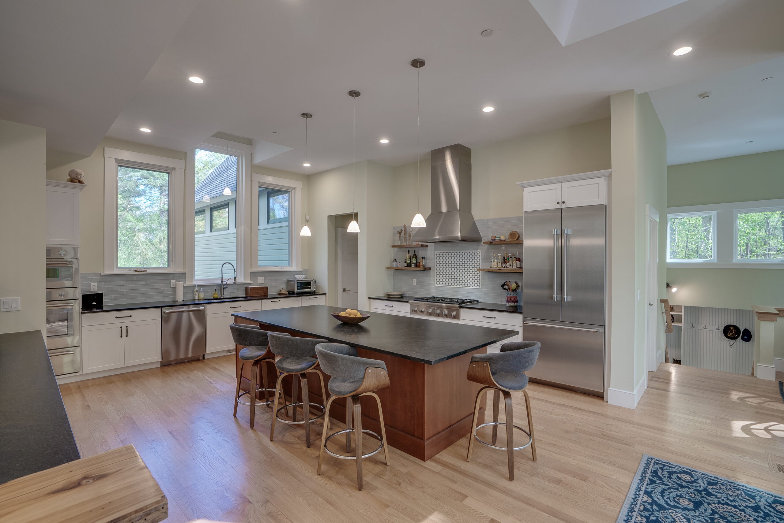 A kitchen island with a dark top and seating for four. The appliances are stainless steel. There are three pendant lights and recessed lighting. Three tall windows sit over the sink in the background. The flooring is wood.