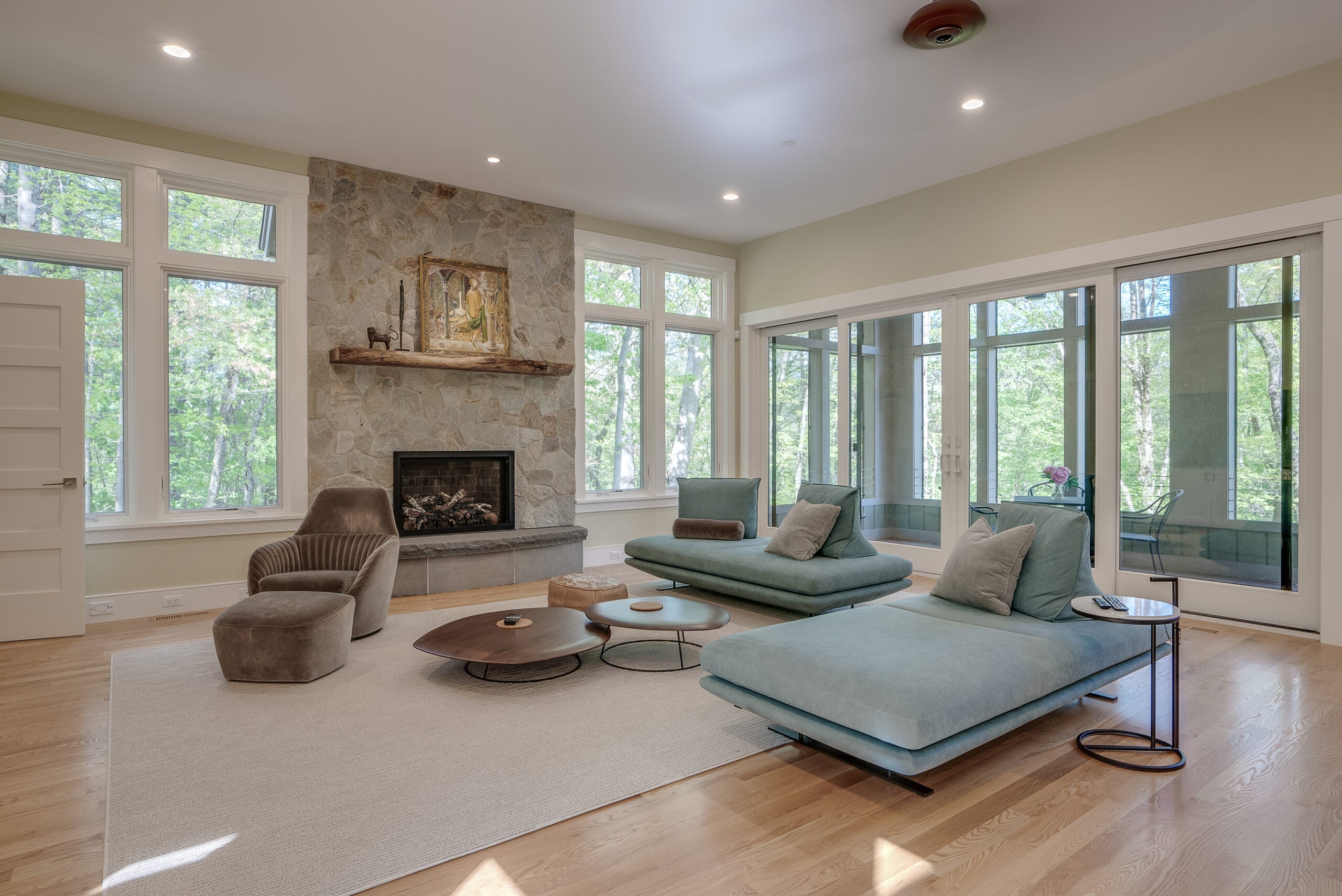 a room with tall windows that extend to the ceiling, hardwood flooring, blue couches with no arms but big pillows, a stone fireplace, a gray armchair with an ottoman, and a rounded, three-tier coffee table.