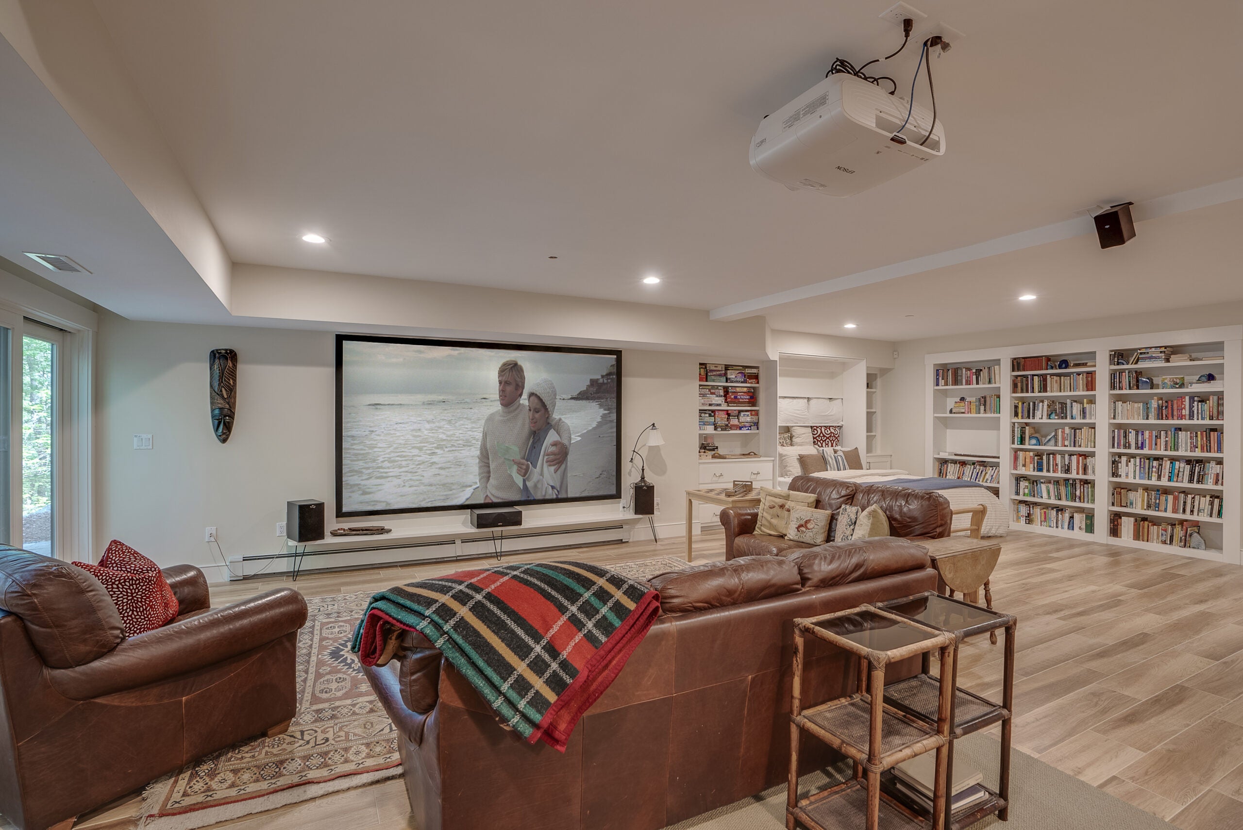 A room with a projector, leather couches and chairs, a big screen, recessed lighting, and white walls lined with bookshelves in a Bedford home.