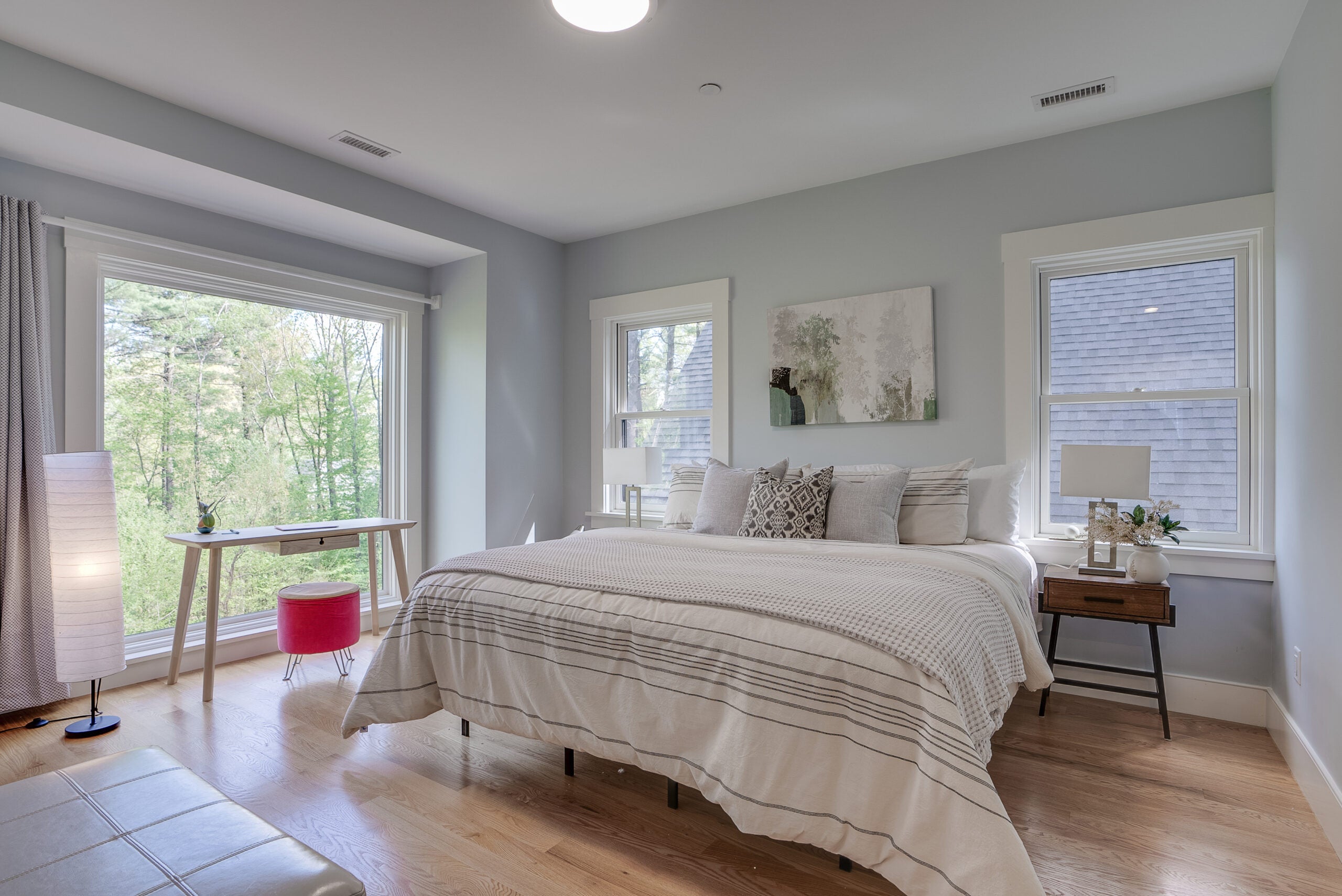 A bedroom with two double-hung windows, gray walls, wood flooring, and a big picture window. A desk is set before the window.