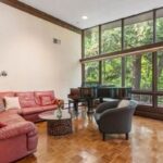 A room with cream-colored walls with no artwork on them. A round pinkish sectional is the focal point. A beam bisects a wall of windows. A grand piano sits before the windows. The flooring is wood and parquet. The coffee table is glass and circular. The photo is for a story on open houses.