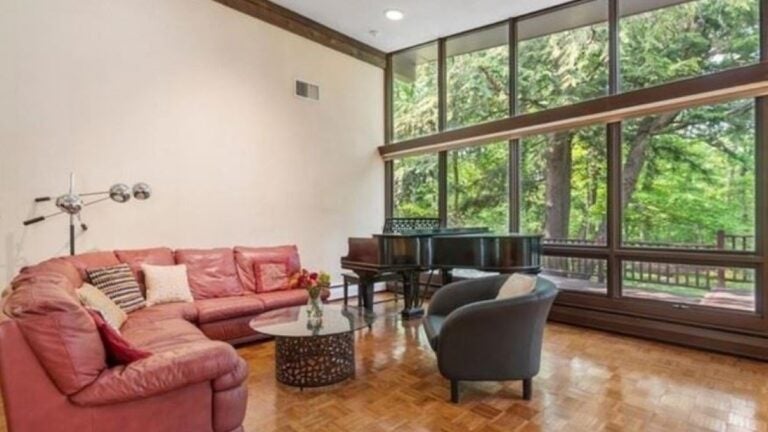 A room with cream-colored walls with no artwork on them. A round pinkish sectional is the focal point. A beam bisects a wall of windows. A grand piano sits before the windows. The flooring is wood and parquet. The coffee table is glass and circular. The photo is for a story on open houses.