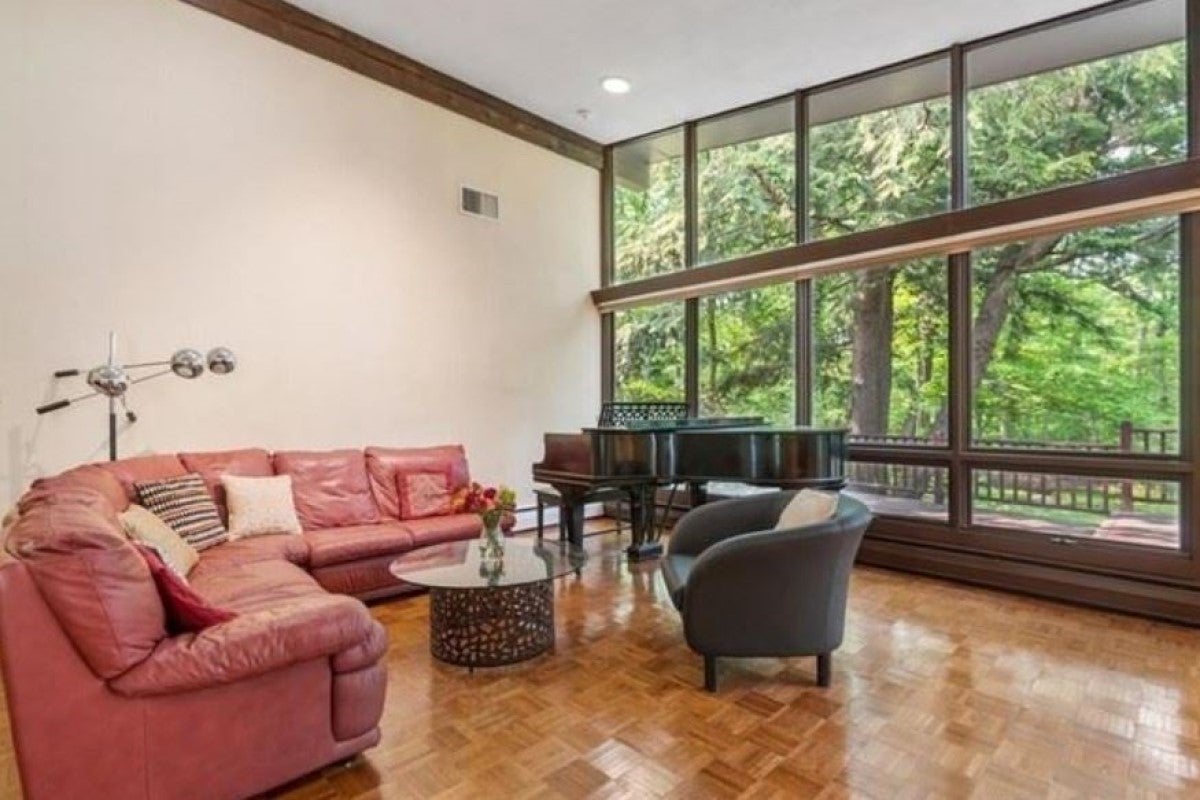 A room with cream-colored walls with no artwork on them. A round pinkish sectional is the focal point. A beam bisects a wall of windows. A grand piano sits before the windows. The flooring is wood and parquet. The coffee table is glass and circular. The photo is for a story on open houses.