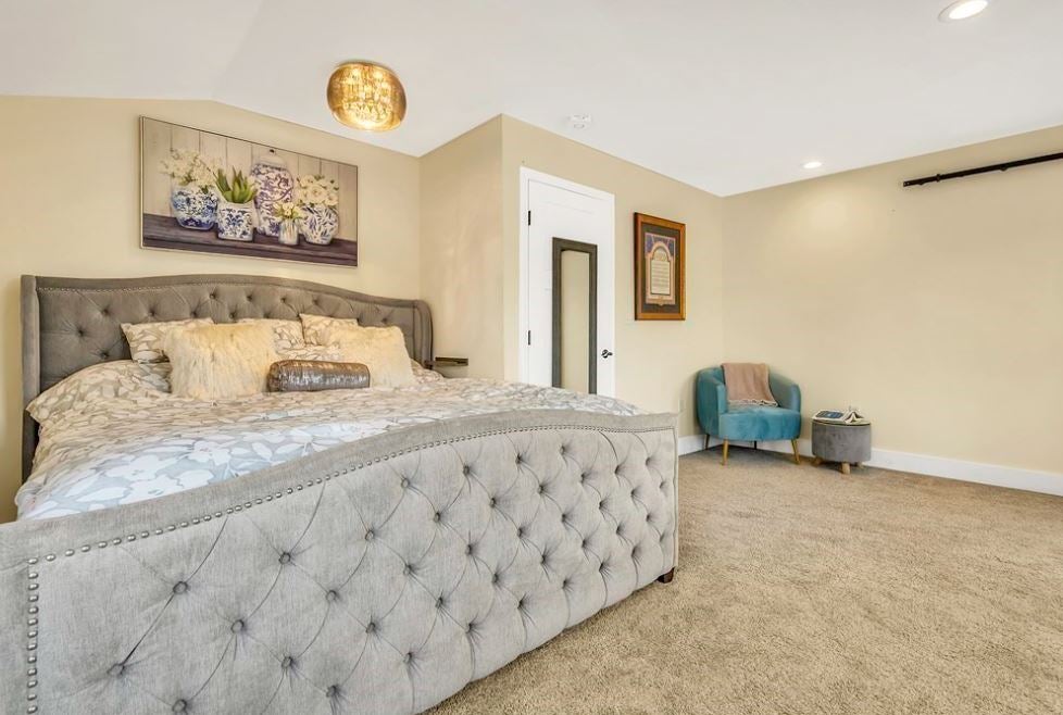 A room with beige walls, a round, bronze light fixture, and a bed with gray, tufted footboard and headboard. The carpeting is beige.