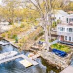A white multi-story home with a deck that spans the width of the home. The home is white. The yard slopes down to a lake, as well as a dock. There's a boat docked. There's also a dock for swimmers. The yard ends in a rock wall. A big, leafless tree bisects the photo.