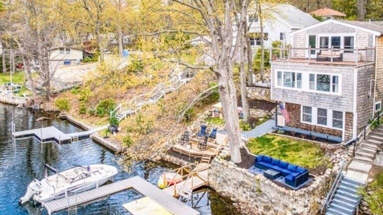 A white multi-story home with a deck that spans the width of the home. The home is white. The yard slopes down to a lake, as well as a dock. There's a boat docked. There's also a dock for swimmers. The yard ends in a rock wall. A big, leafless tree bisects the photo.