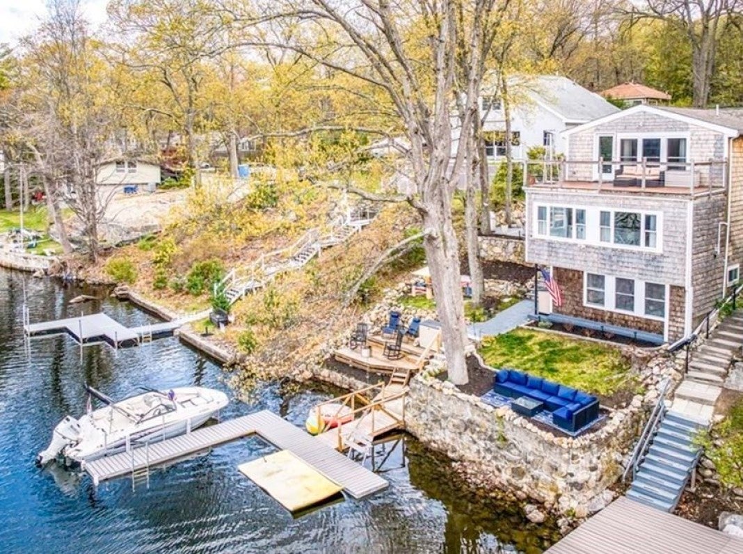 A white multi-story home with a deck that spans the width of the home. The home is white. The yard slopes down to a lake, as well as a dock. There's a boat docked. There's also a dock for swimmers. The yard ends in a rock wall. A big, leafless tree bisects the photo.