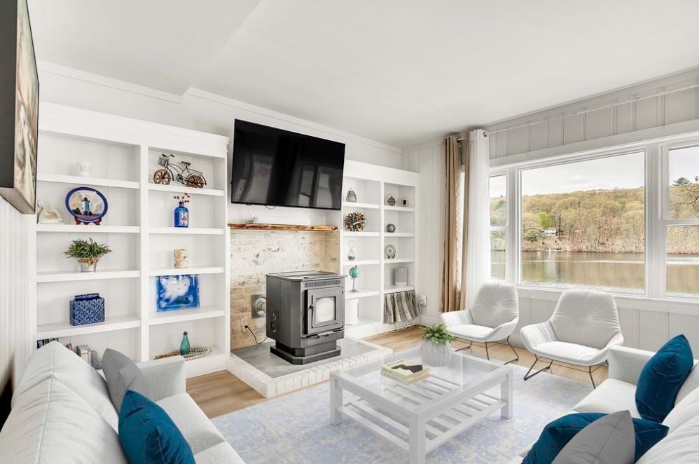 A room with light-colored walls, a woodstove and TV flanked by white built-in cabinetry and bookshelves, two white Eames chairs, a white couch with medium blue throw pillows, and a wide muntin-less window overlooking a lake. A square white coffee table with a glass top holds center court.