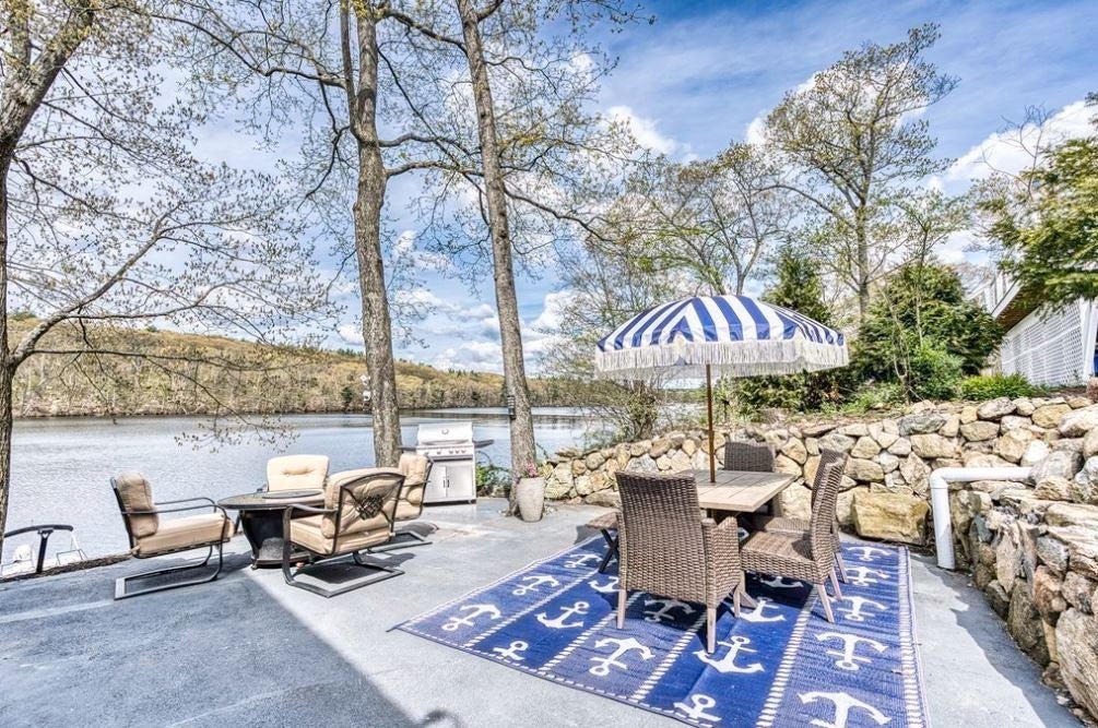 A patio along the water in the early spring with wicker chairs, a table with a blue-and-white striped umbrella, and chairs around a firepit.