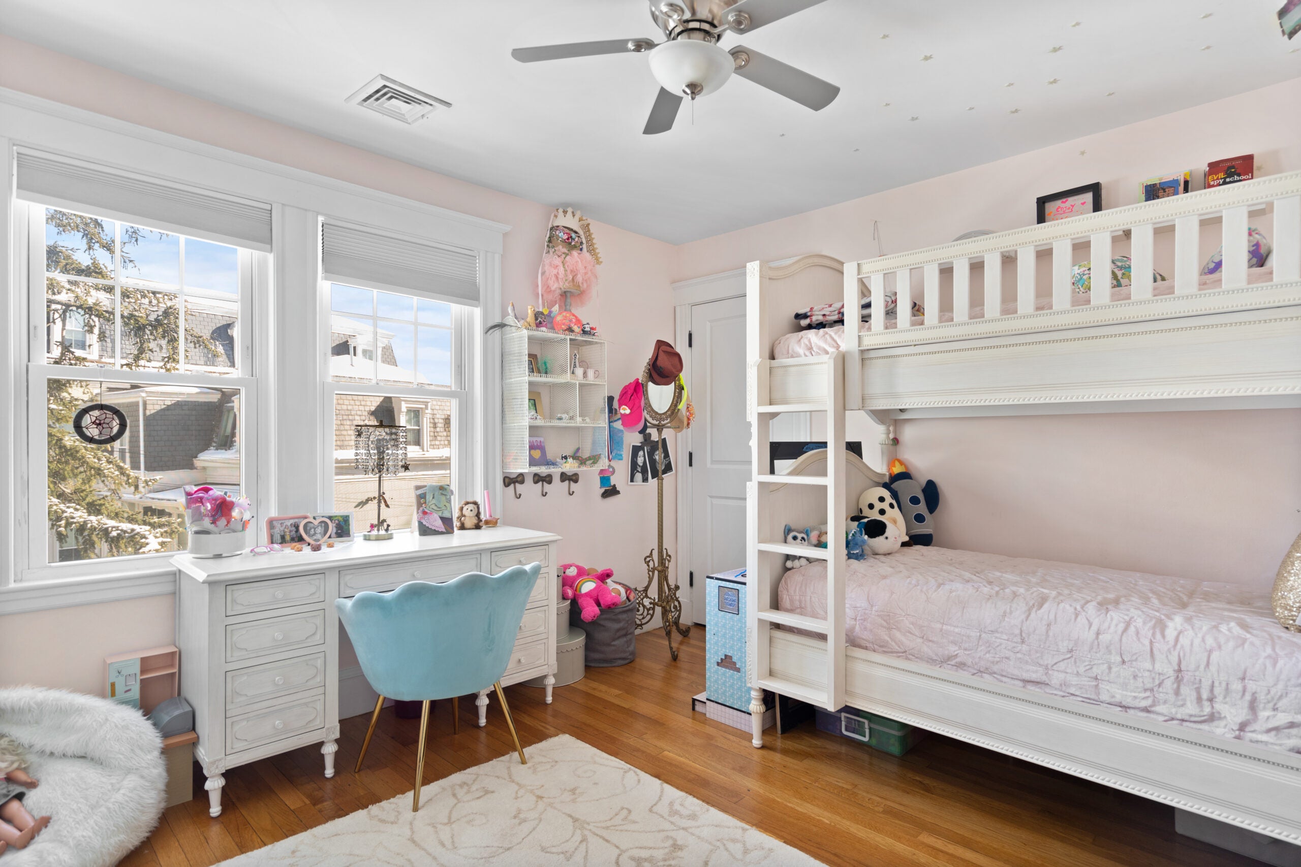A room in a Newton condo with white trim, light-pink walls, a white desk with a blue tulip chair, a white bunkbed with flowery pink bedding, wood flooring, a light-colored area rug, a beanbag chair, and a ceiling fan.