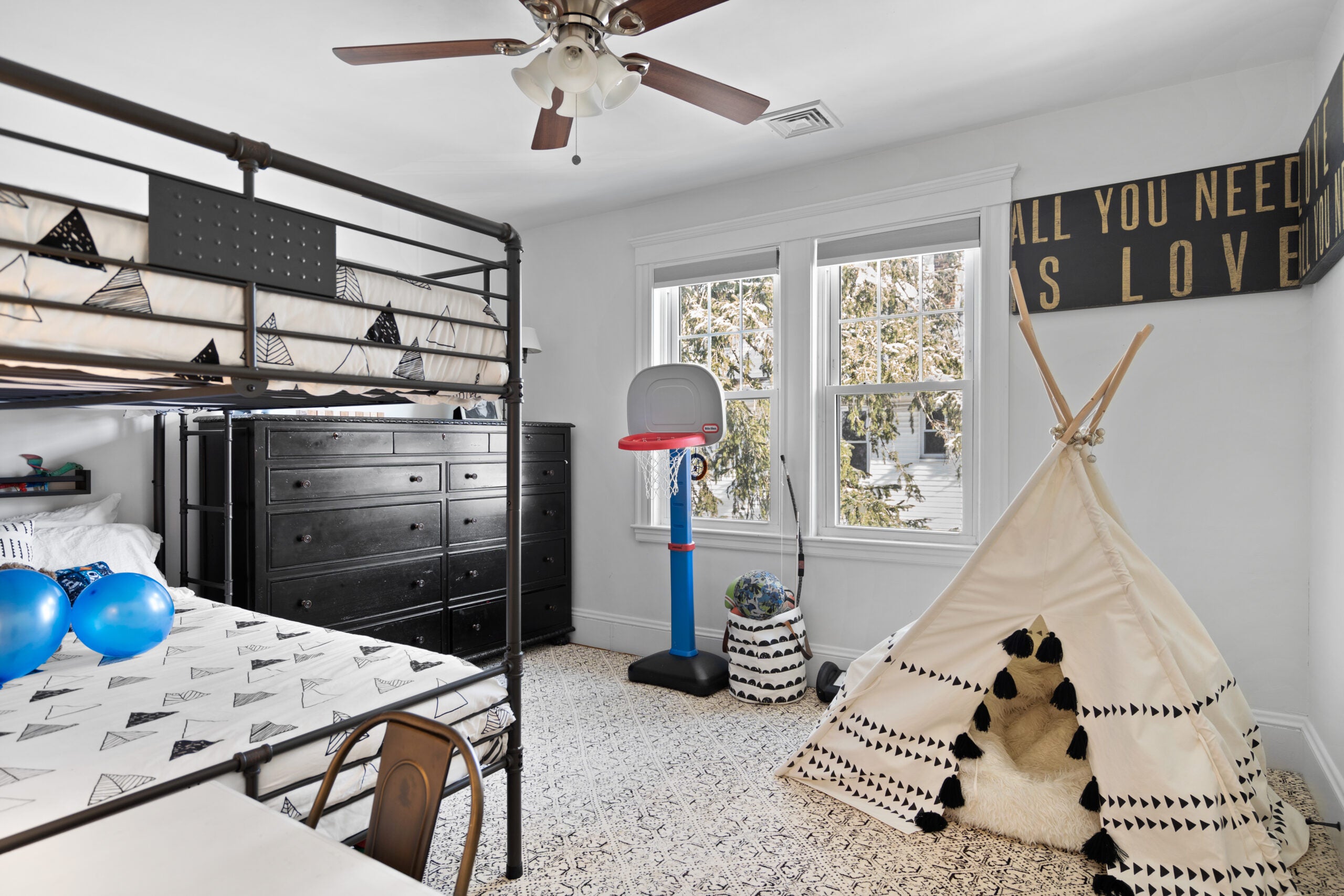 A child's bedroom with gray walls, white trim, a teepee, a sign that reads "All you need is love" in gold allcaps on a black background, a ceiling fan, wood flooring, an area rug, a black dresser, and a blue plastic basketball hoop.