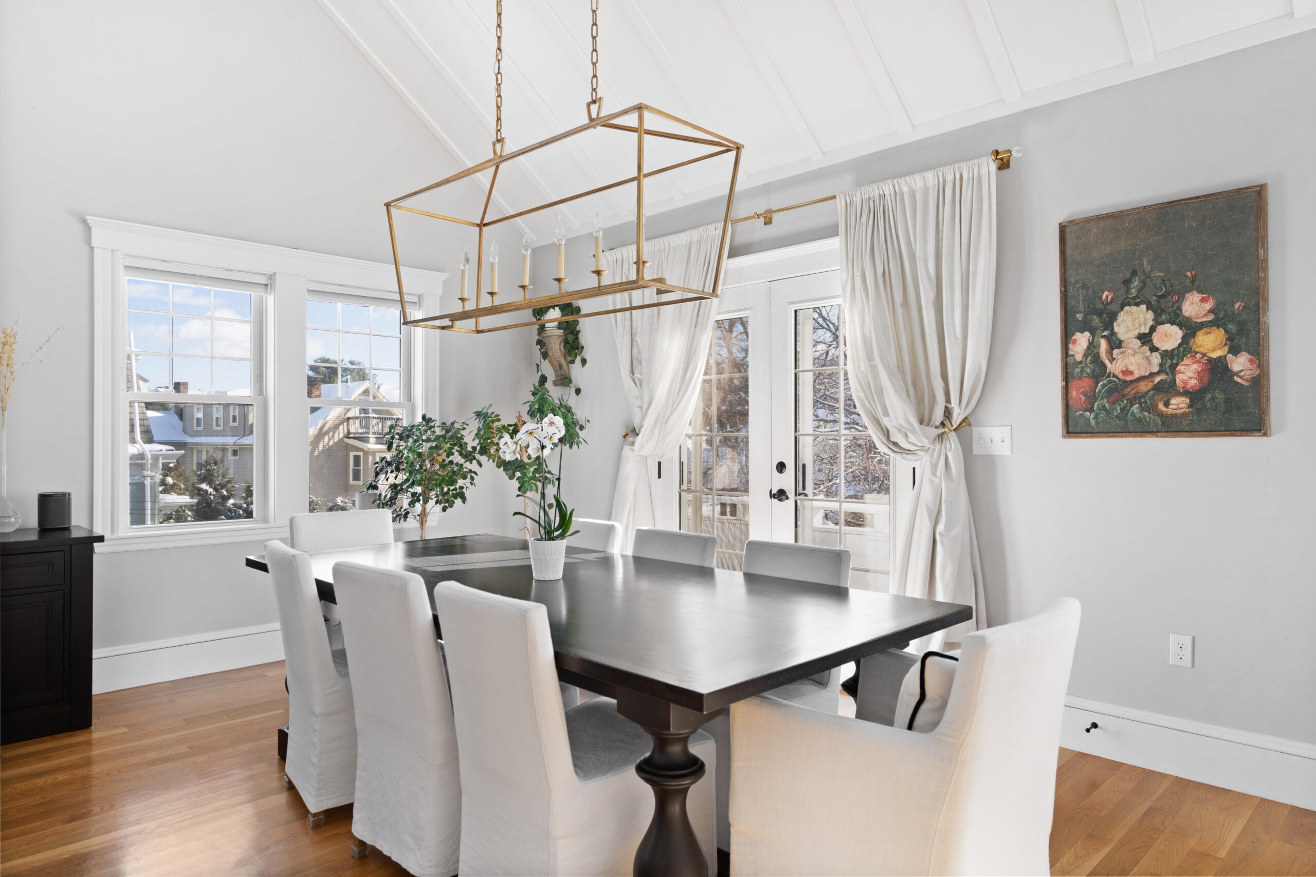 A room in a Newton condo with gray walls, a vaulted ceiling, white trim, two paired windows, wood flooring, French doors to a balcony, a boxy and brass chandelier, and a long and dark dining table paired with white upholstered chairs.
