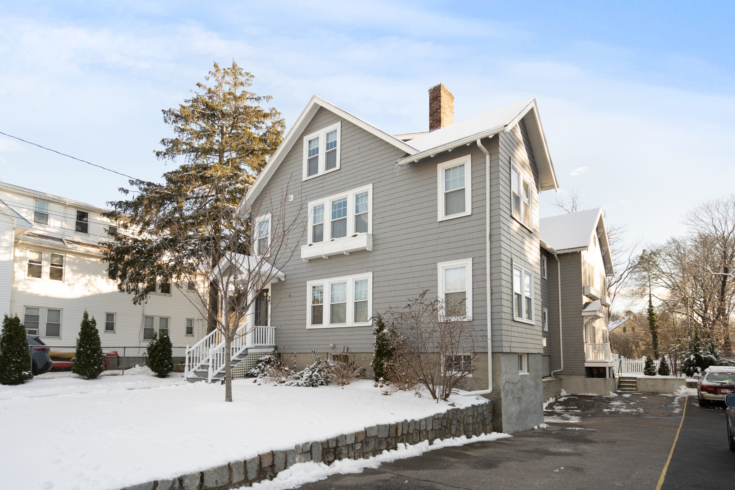 A gray multifamily home in Newton with white trim, 12 windows, a lone tree out front, snow on the group, blue skies, and a driveway running front to back in the foreground.