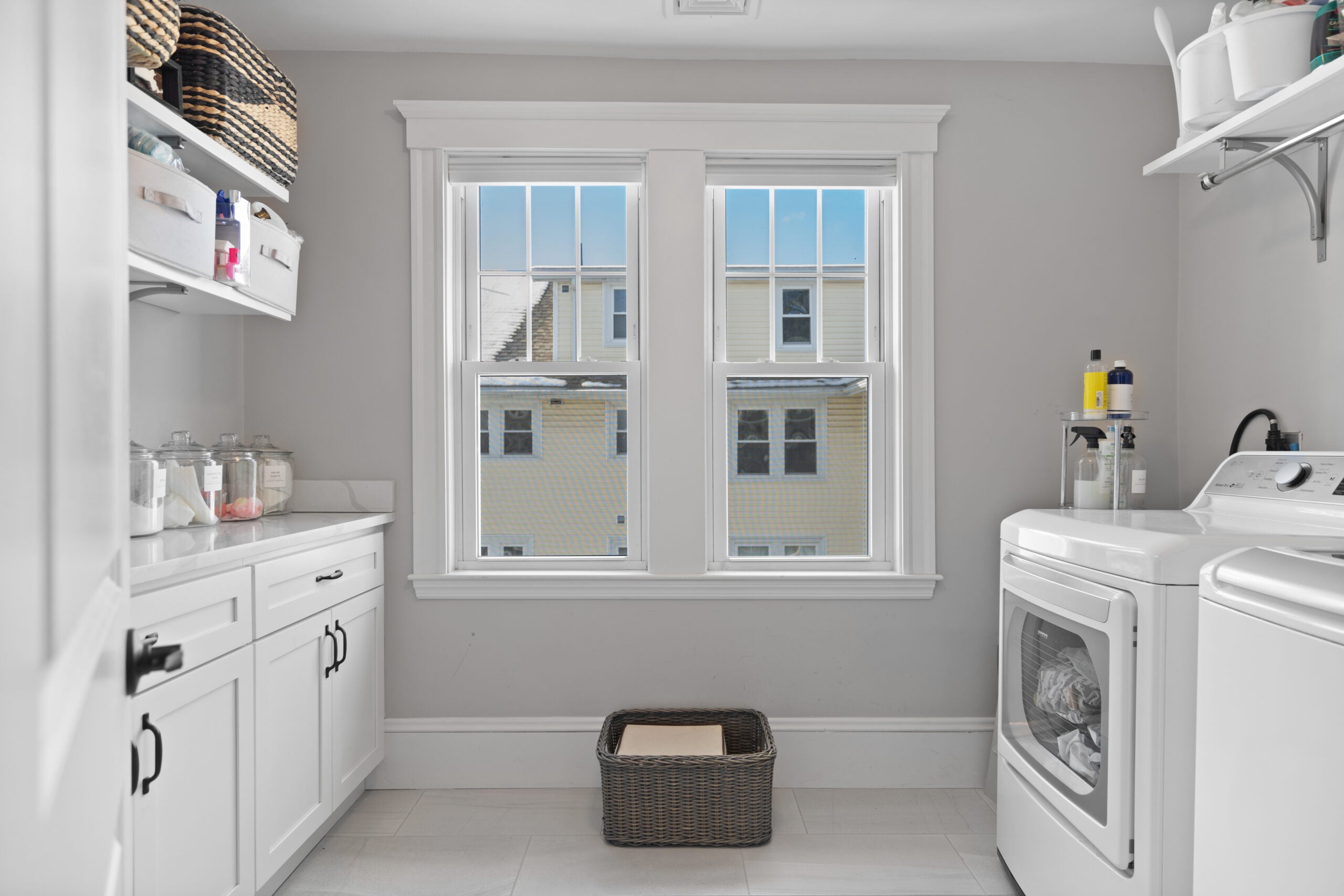 A laundry room with white cabinetry (upper and lower) on one side, a pair of windows in the back, gray walls, and on the far right: a front-load dryer next to a top-load washer. Both sit under a white shelf with a drying rack.