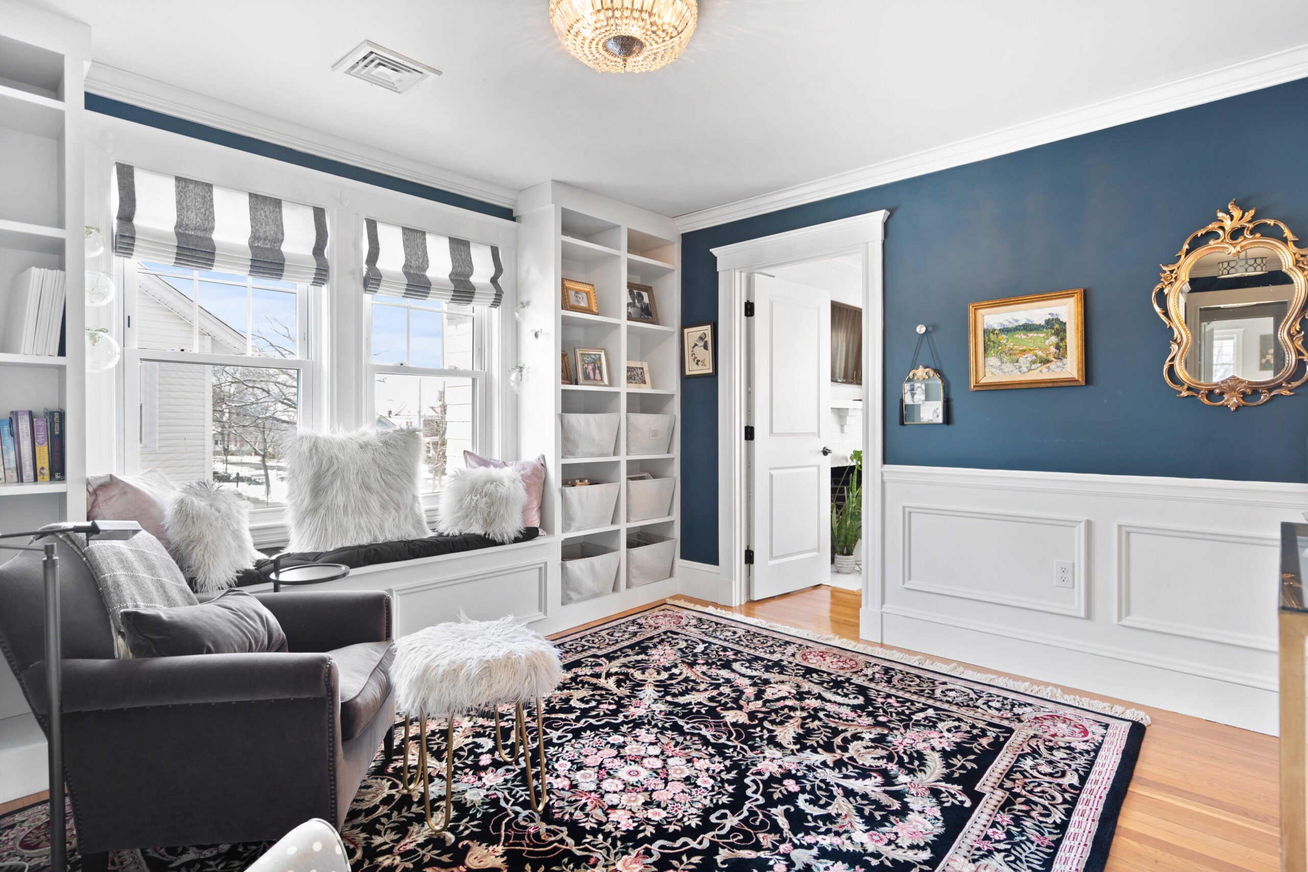 A room with peacock blue walls, white picture-frame wainscoting, a windows seat with a blue cushion and white pillows, wood flooring with an Oriental rug, gray-and-white vertically striped Roman shades, and white built-in bookshelves. There's also a dark gray arm chair with a fuzzy white ottoman.