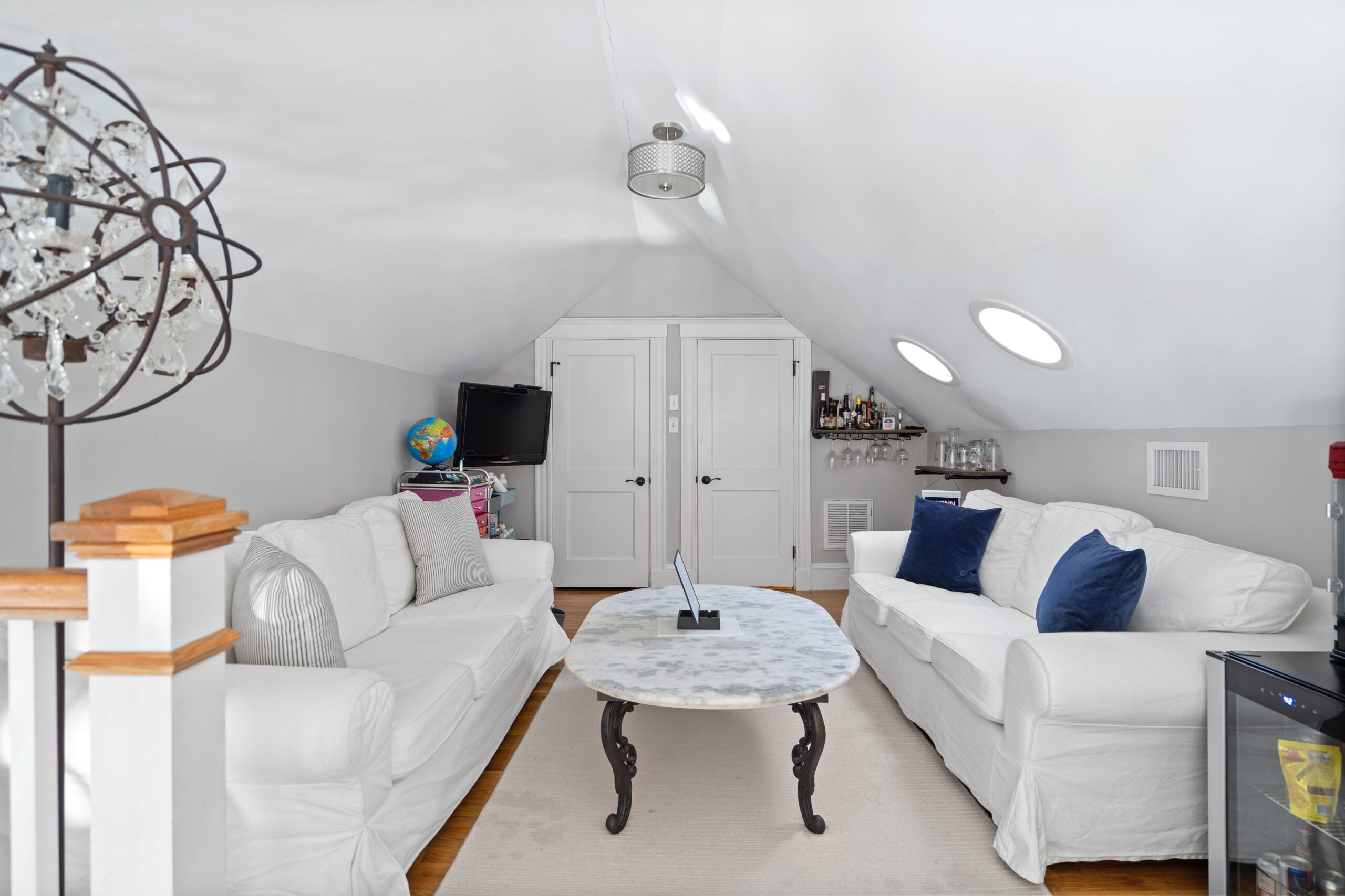 A loft area with a pair of white couches that flank an overall coffee table with a marble top. The room has recessed lights, wood flooring, a gray area rug, and blue and gray throw pillows. There are two doors at the back of the room.