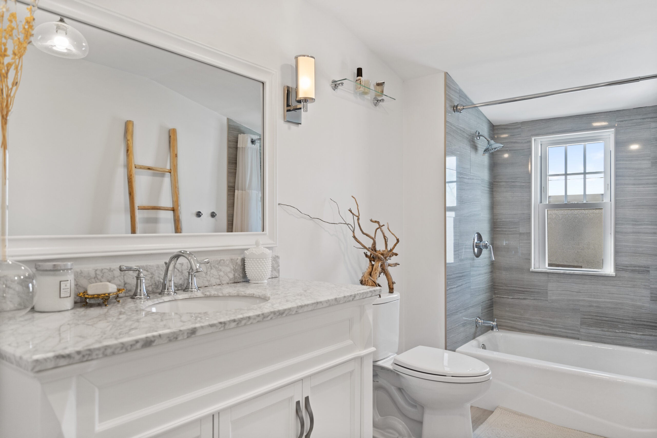 A look at the main bathroom, which has white walls, sconces, a rectangular mirror framed in white to match the cabinetry, and a tub-shower combination. The tile around the shower is gray.