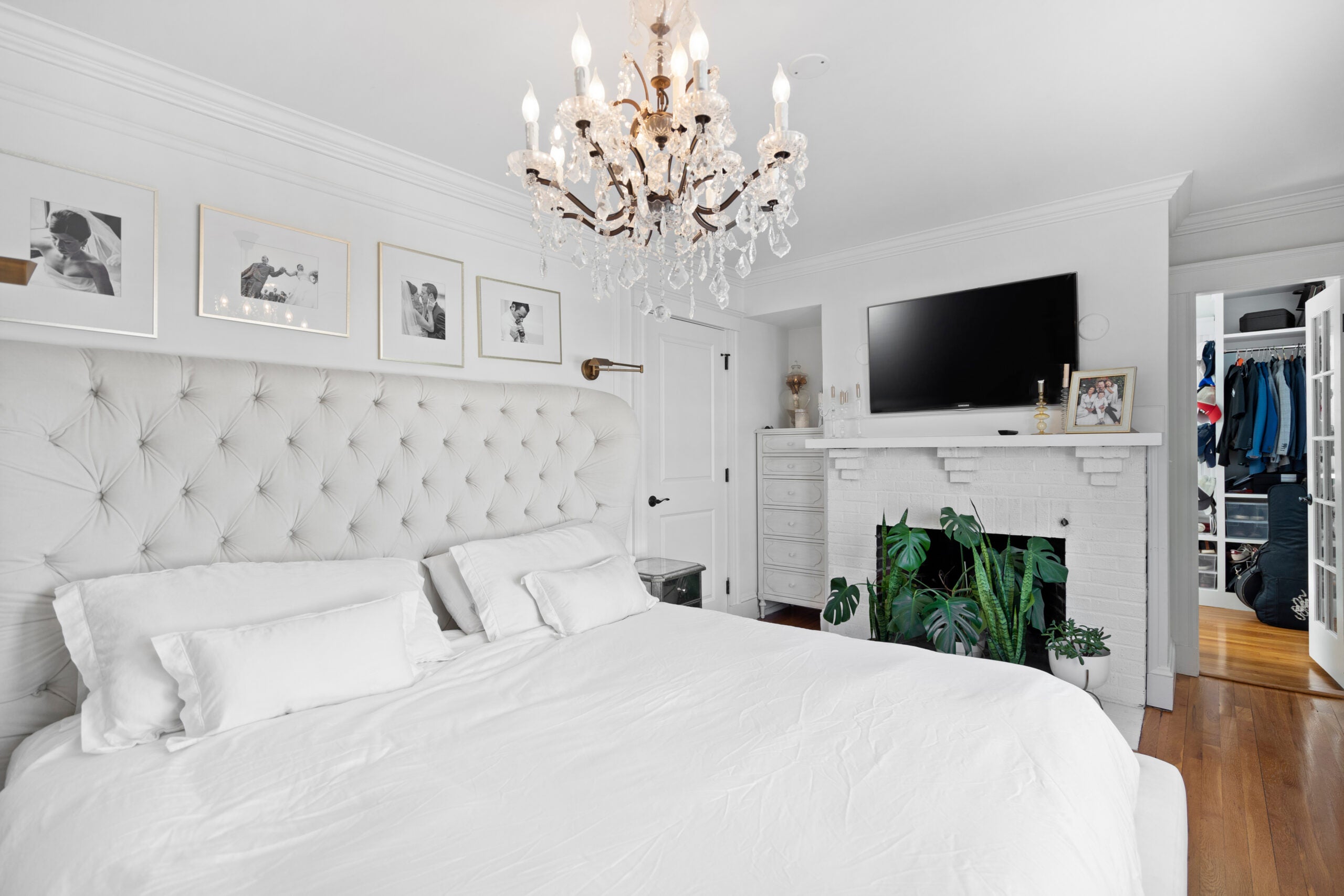 A room in which nearly everything is white, from the walls and brick fireplace to the bedding. The cushiony headboard is gray. The flooring is wood. There is a crystal chandelier with candle-like lights over the bed. One can see the walk-in closet in the back.