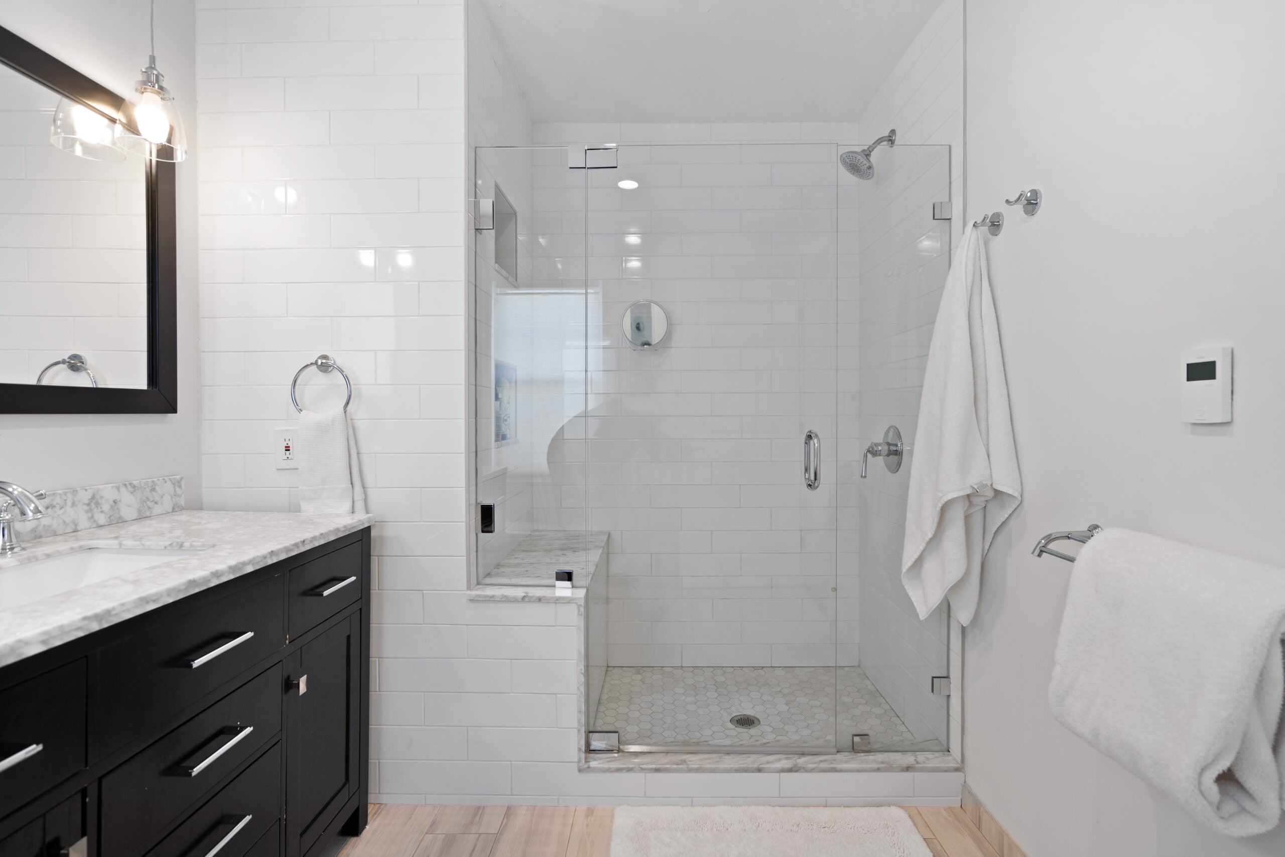 A long view of the room, with an espresso-colored vanity on the left under a rectangular mirror of the same hue. the vanity is topped with a marbled quartz. The wall next to and in the shower is subway tile. The shower has a clear glass door. The flooring is a light-colored tile. The walls are white. There is a towel rack on the far right.