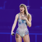 Taylor Swift laughs on stage at Gillette Stadium in a sparkly body suit. The background is tinted purple.