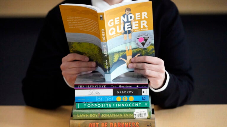 Person reading "Gender Queer" behind a stack of books that have been the subject of complaints
