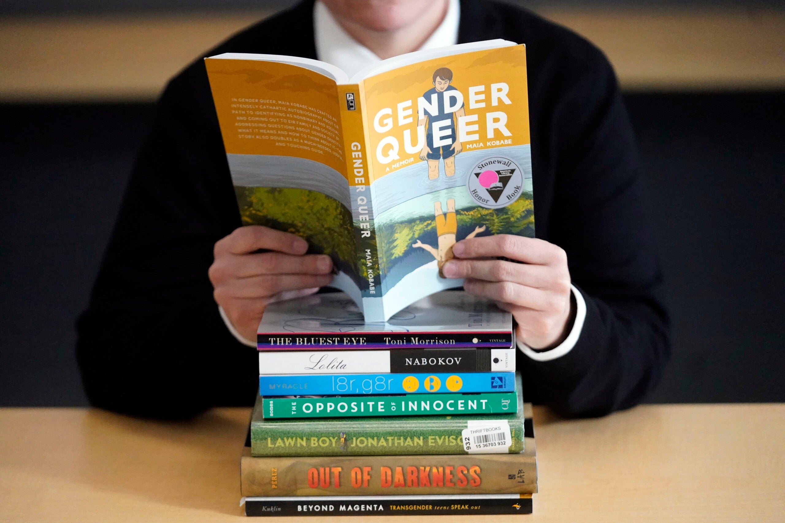 Person reading "Gender Queer" behind a stack of books that have been the subject of complaints