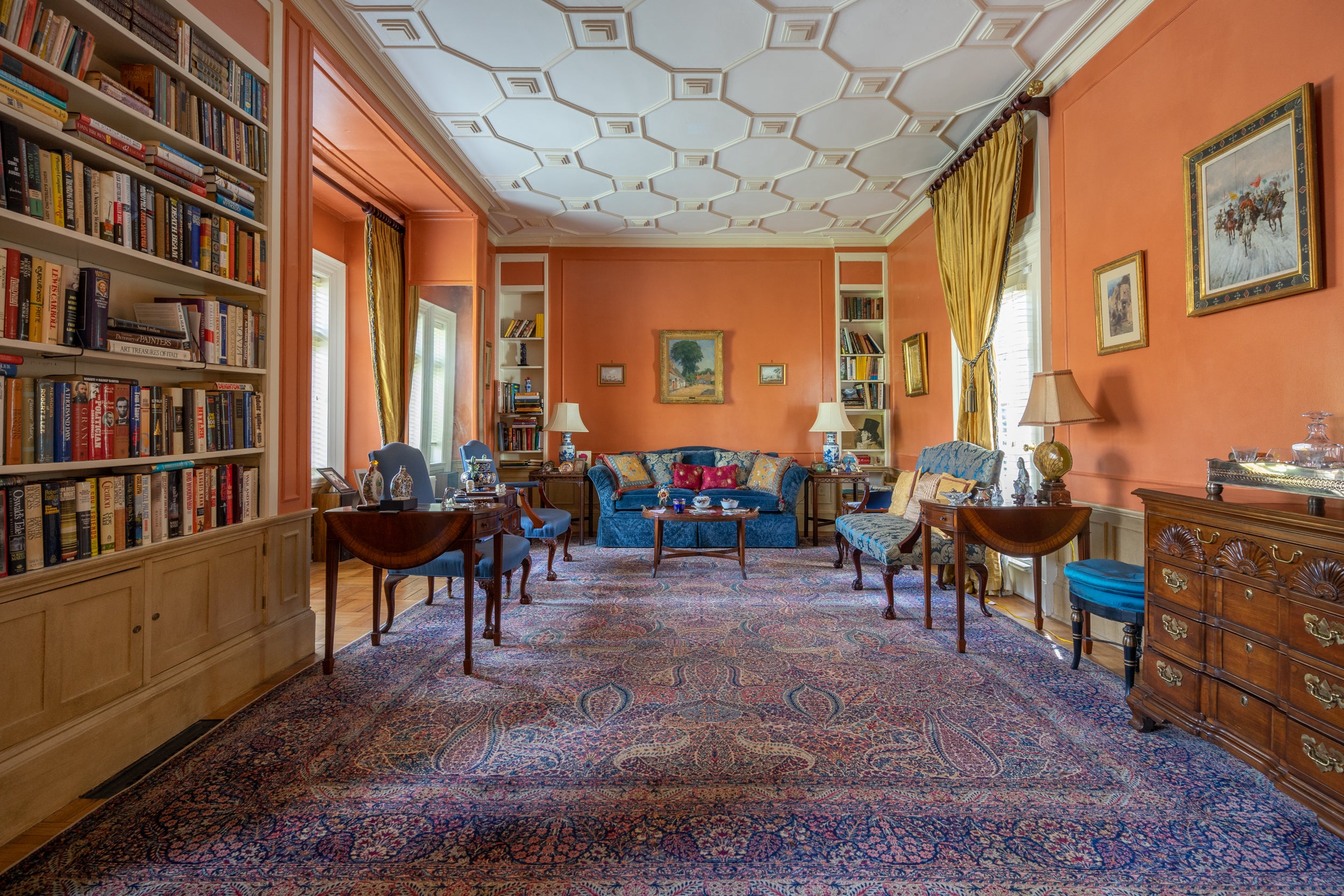 The library has intricate ceiling moulding and orange walls, picture windows, and built-in bookshelves.