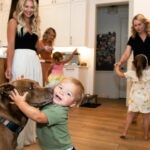 A child plays with a dog in the foreground, while a mother dances with her daughter in the background, another mother helps a child by the kitchen counter, and another mother watches the toddler and dog play.