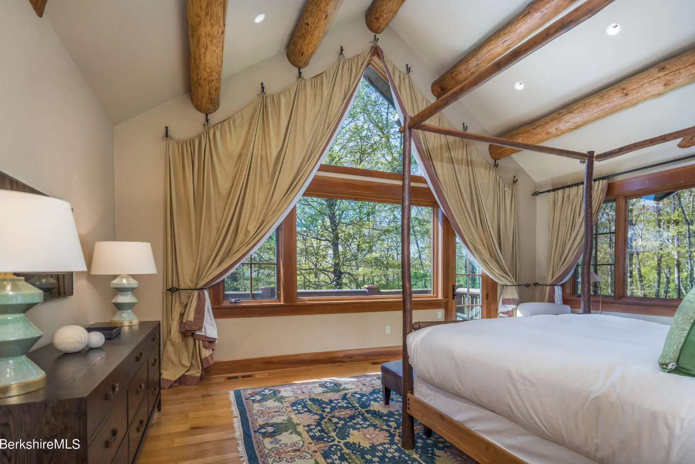 Primary bedroom with vaulted ceilings, picture windows with floor-to-ceiling curtains, hardwood floors, and exposed wood beams.