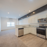 Open floor plan kitchen and living room. The kitchen has white Shaker-style cabinets and stainless steel appliances, and the living area has white walls, single-hung windows, and beige carpeting.