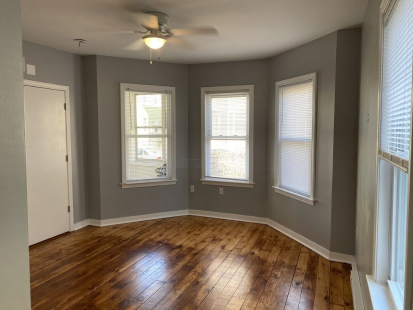 Living area with dark gray walls and single-hung bay windows. The room has hardwood floors and a ceiling fan.