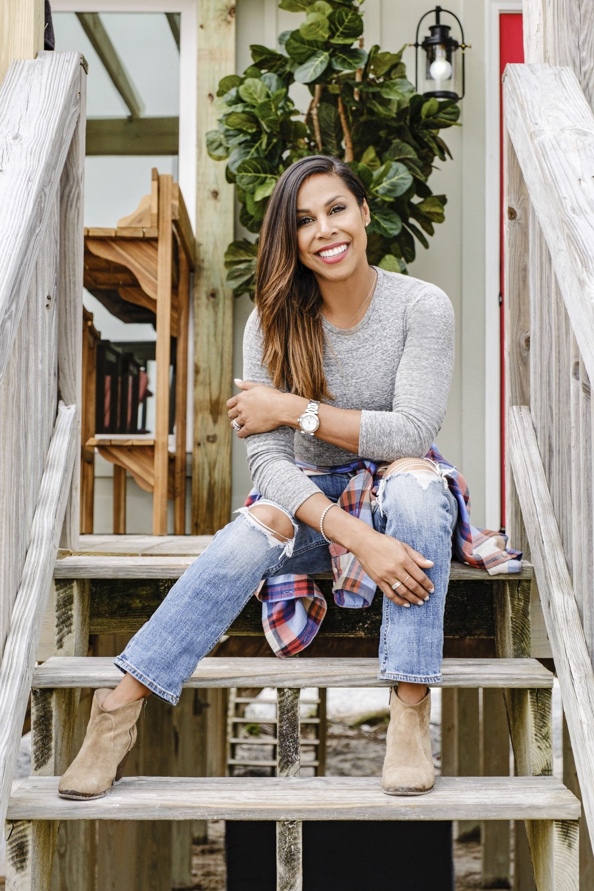 Taniya Nayak of HGTV's Battle on the Beach. She is wearing a heather cray long-sleeved shirt, has a red plaid flannel tied around her waist, and is wearing blue jeans while sitting on steps.