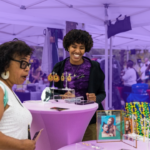 Two women look at tables of crafts at the Black Owned Bos. market in Seaport under tents. The background is tinted purple.