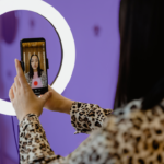 A woman holds up her phone in selfie-mode recording a video in front of a ring light. The background is tinted purple.