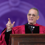 Tom Hanks stands at a brown podium in a red doctorate graduation gown giving a speech at Harvard's commencement. The background of the image is tinted purple.