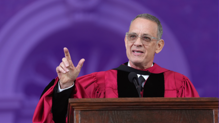 Tom Hanks stands at a brown podium in a red doctorate graduation gown giving a speech at Harvard's commencement. The background of the image is tinted purple.