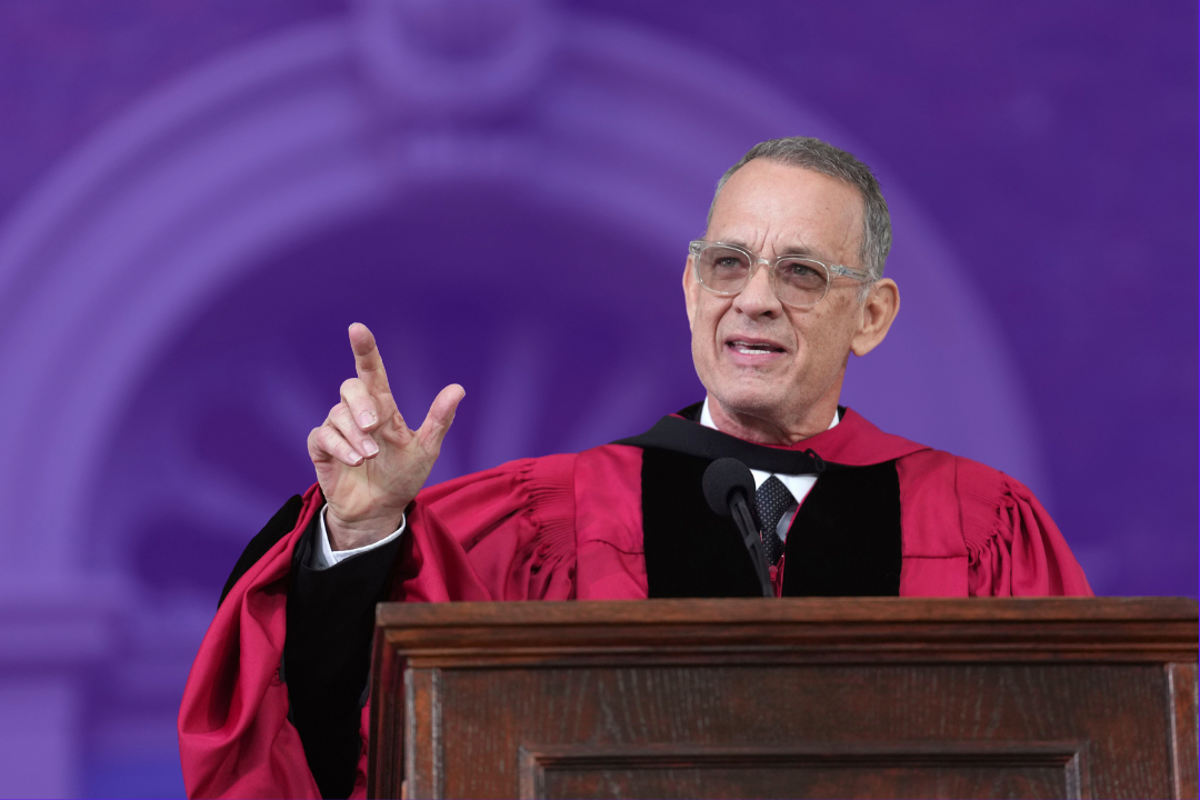 Tom Hanks stands at a brown podium in a red doctorate graduation gown giving a speech at Harvard's commencement. The background of the image is tinted purple.