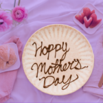 A plate with "Happy Mother's Day" written on it in chocolate sauce sits on a bed spread and is surrounded by flowers, a croissant, and strawberries. The background is tinted purple.