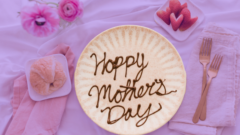 A plate with "Happy Mother's Day" written on it in chocolate sauce sits on a bed spread and is surrounded by flowers, a croissant, and strawberries. The background is tinted purple.