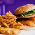 A fried fish sandwich on a sesame bun is on a white plate with a pile of waffle fries. The background is tinted purple.